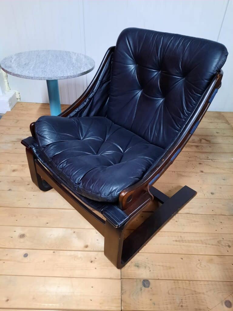 Een zwarte leren loungestoel met gecapitonneerde kussens en een donkerhouten frame staat op een lichte houten vloer naast een kleine ronde tafel met een grijs marmeren blad en een blauwe voet.